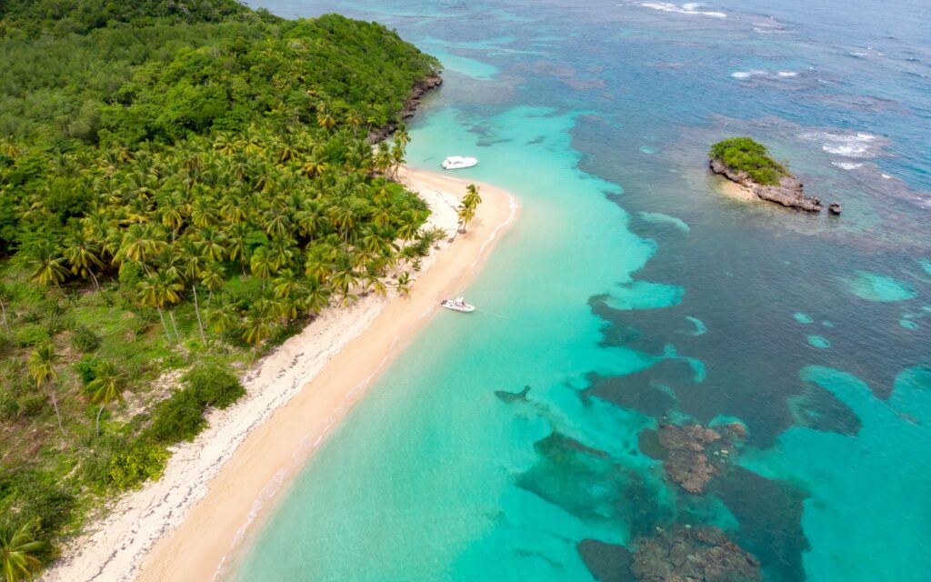 Aerial view of a pristine Caribbean beach with turquoise waters, lush palm trees, and anchored boats, representing the vibrant environment for solar panel maintenance in the Caribbean.